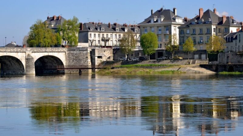Vue d’un pont en pierre et de maisons traditionnelles se reflétant dans une rivière calme, par temps ensoleillé.