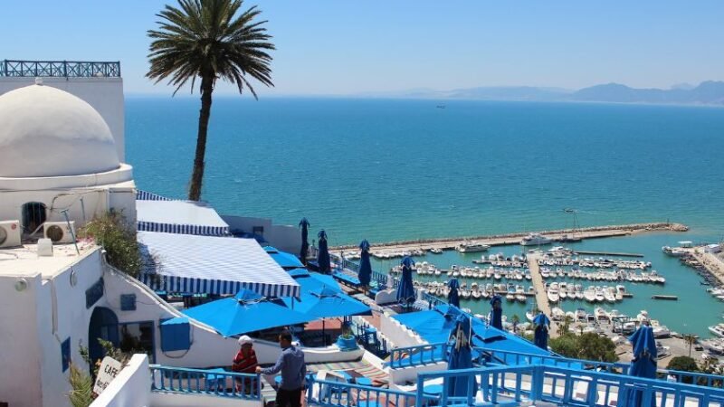 Vue d’un café en terrasse blanc et bleu surplombant un port de plaisance et la mer Méditerranée.
