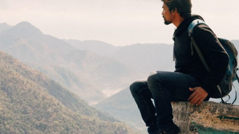 Homme assis au bord d’une falaise avec un sac à dos, contemplant une vue panoramique sur des montagnes verdoyantes sous un ciel nuageux.