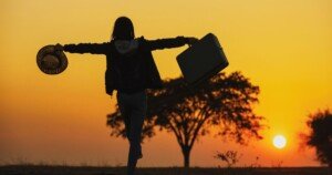 Silhouette d’une voyageuse levant les bras face au coucher du soleil, portant une valise et un chapeau, symbolisant la liberté, l’aventure et le tourisme conscient.