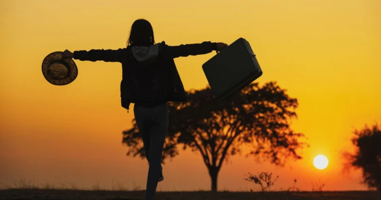 Silhouette d’une voyageuse levant les bras face au coucher du soleil, portant une valise et un chapeau, symbolisant la liberté, l’aventure et le tourisme conscient.