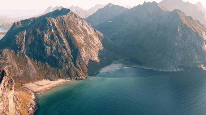 Vue aérienne d’une côte sauvage avec plages isolées et hautes montagnes plongeant dans une mer calme, sous une lumière douce.
