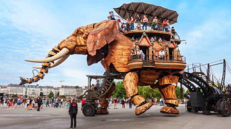 Éléphant mécanique géant transportant des visiteurs lors d’une attraction artistique en plein air, dans une ville européenne animée.