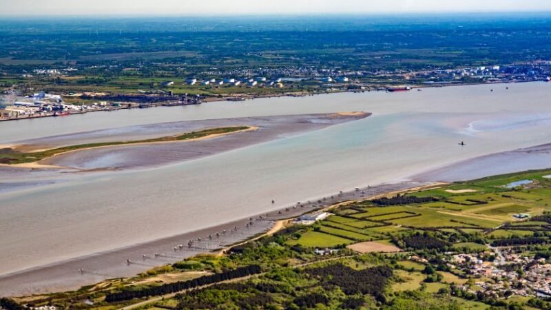 Vue aérienne d’un large estuaire avec zones humides, champs agricoles et ville côtière à l’horizon.