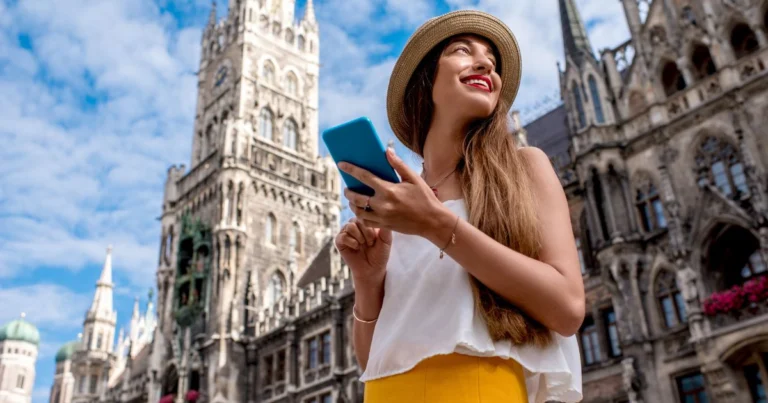 Jeune femme souriante avec un smartphone explorant le centre historique de Munich, devant l’hôtel de ville gothique, symbole de tourisme urbain connecté et curieux.