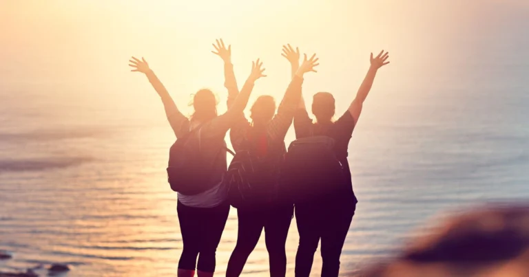 Trois voyageuses avec sacs à dos levant les bras face au coucher de soleil sur la mer, symbolisant la liberté, la sororité et l’esprit du voyage responsable et solidaire.