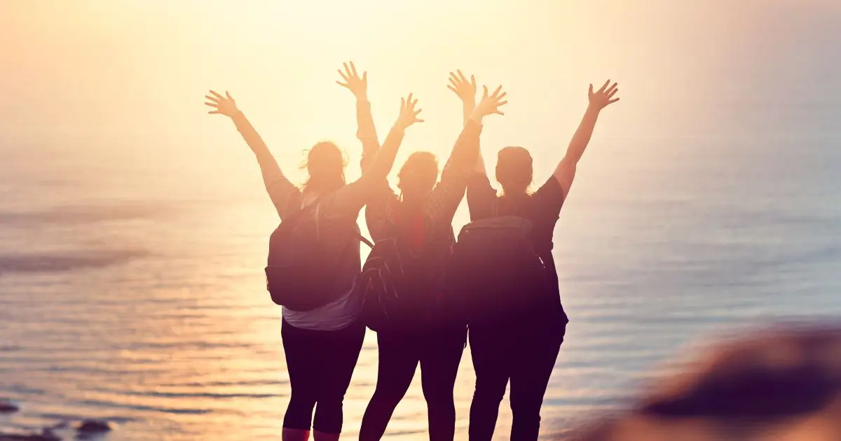 Trois voyageuses avec sacs à dos levant les bras face au coucher de soleil sur la mer, symbolisant la liberté, la sororité et l’esprit du voyage responsable et solidaire.