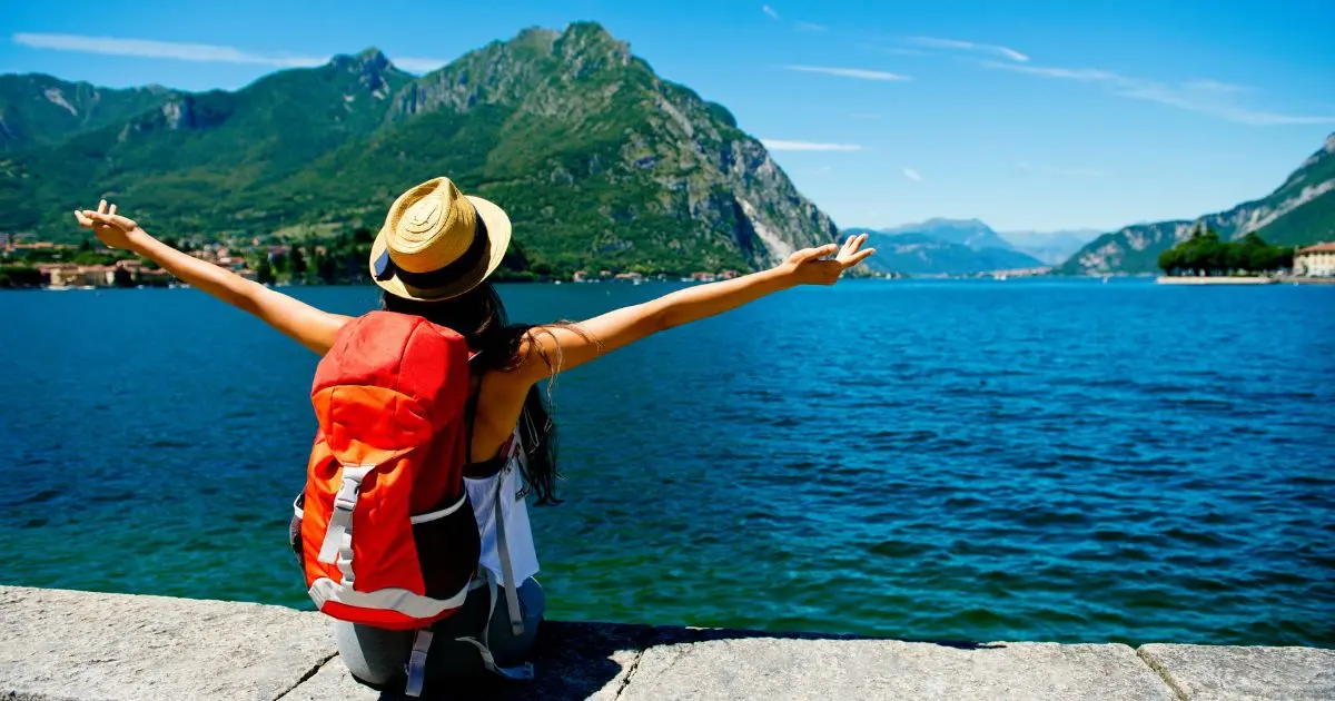 Voyageuse assise face à un lac entouré de montagnes verdoyantes, les bras levés en signe de liberté, illustrant un tourisme conscient et harmonieux avec la nature.