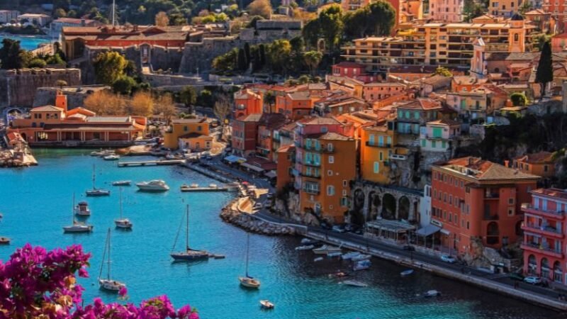 Village méditerranéen coloré en bord de mer avec bateaux amarrés, eau turquoise et collines verdoyantes en arrière-plan.