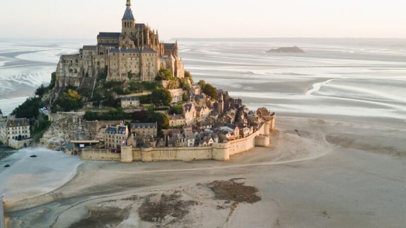 Vue aérienne du Mont-Saint-Michel avec ses remparts et son abbaye au lever du soleil, entouré de vastes étendues de sable.