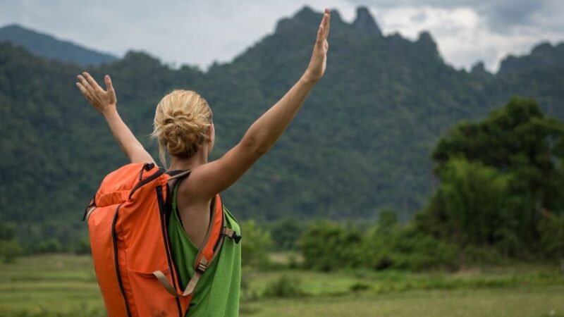 Femme de dos avec un sac à dos orange, bras levés vers le ciel, admirant un paysage naturel de montagne et de forêt sous un ciel nuageux.