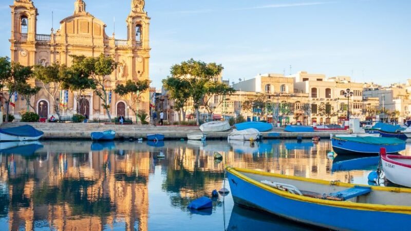 Port méditerranéen avec bateaux colorés amarrés, reflet d’une église historique et bâtiments traditionnels sur l’eau calme.
