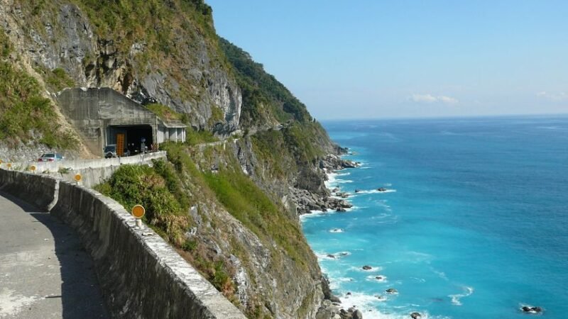 Route côtière sinueuse longeant des falaises rocheuses avec tunnel, vue panoramique sur une mer turquoise et ciel dégagé.
