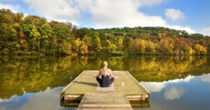 Femme en méditation assise sur un ponton en bois, face à un lac calme entouré de forêts colorées en automne.