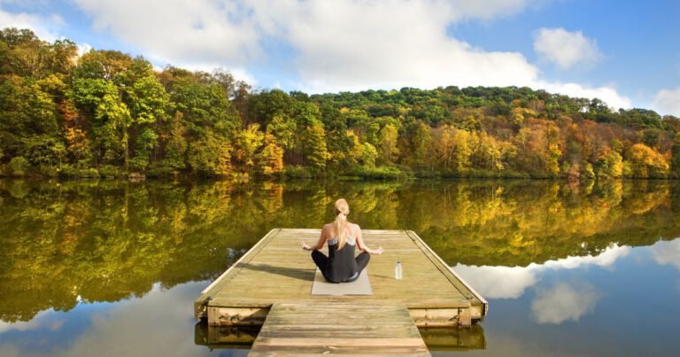 Femme en méditation assise sur un ponton en bois, face à un lac calme entouré de forêts colorées en automne.