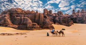 Caravanes de dromadaires traversant un désert rocheux sous un ciel nuageux.