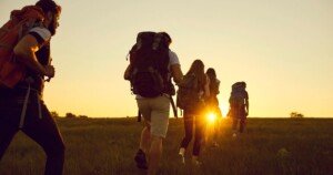 Groupe de randonneurs marchant en file vers le soleil couchant à travers une prairie, symbolisant l’aventure collective, le slow travel et le respect de la nature.