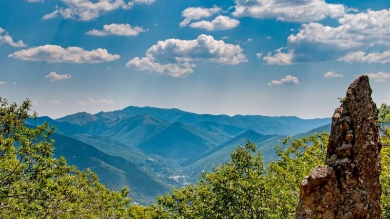 Vue panoramique sur des montagnes verdoyantes sous un ciel bleu parsemé de nuages, avec un rocher en premier plan.