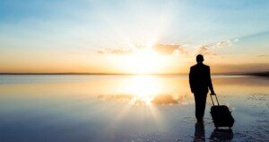 Homme en costume avec valise contemplant un lever de soleil sur une étendue d’eau calme, symbolisant le voyage intérieur, la transition et l’impact du voyageur moderne.