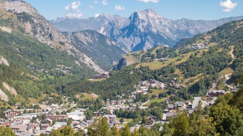 Village de montagne niché dans une vallée verdoyante, entouré de sommets alpins majestueux sous un ciel bleu clair.