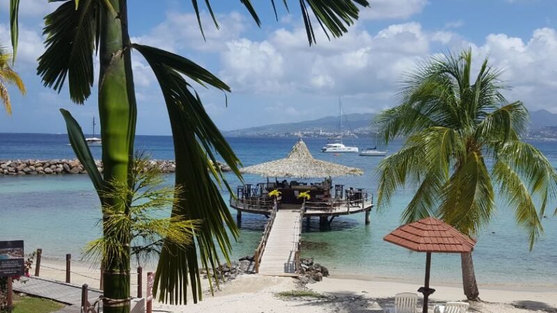 Plage tropicale avec palmiers et ponton en bois menant à un restaurant sur l’eau, vue sur mer turquoise et voiliers.