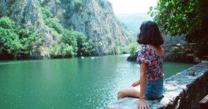 Jeune femme assise au bord d’une rivière entourée de montagnes verdoyantes.