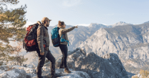 Couple de randonneurs avec sacs à dos observant les montagnes depuis un sommet rocheux, symbole d’aventure, de tourisme responsable et de connexion à la nature.