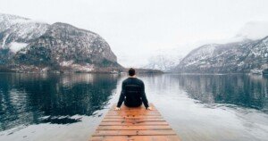 Homme assis au bout d’un ponton en bois, contemplant un lac paisible entouré de montagnes enneigées, symbole de reconnexion à la nature et de tourisme introspectif.