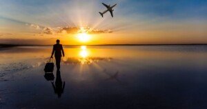 Voyageur en costume marchant au bord de l’eau au coucher du soleil, valise à la main, tandis qu’un avion décolle dans le ciel doré.