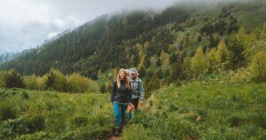 Couple de randonneurs marchant sur un sentier de montagne verdoyant, entouré de forêts de conifères sous un ciel brumeux, dans un paysage naturel propice au tourisme écologique et aux escapades en pleine nature.