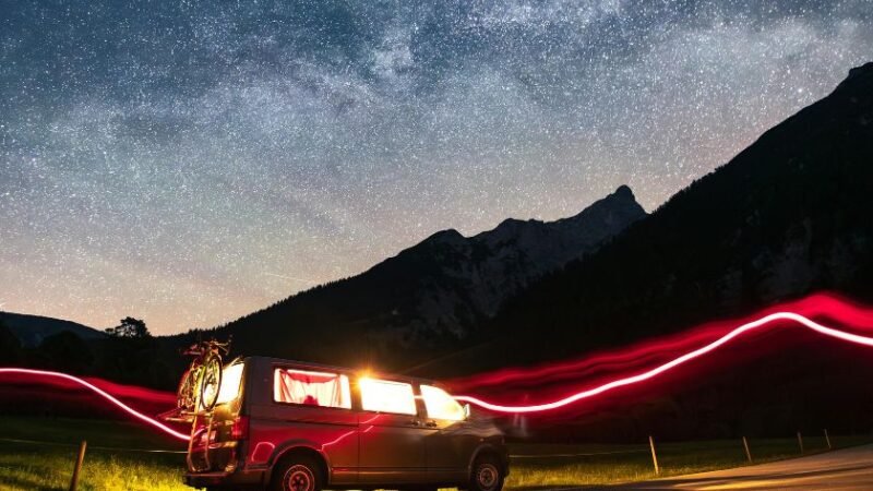 Van aménagé stationné sous un ciel étoilé en montagne, lumière intérieure chaleureuse et traces lumineuses rouges en mouvement.
