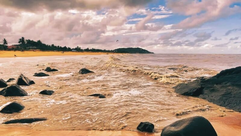 Plage dorée bordée de rochers noirs sous un ciel nuageux, avec des vagues douces et des palmiers à l’horizon.