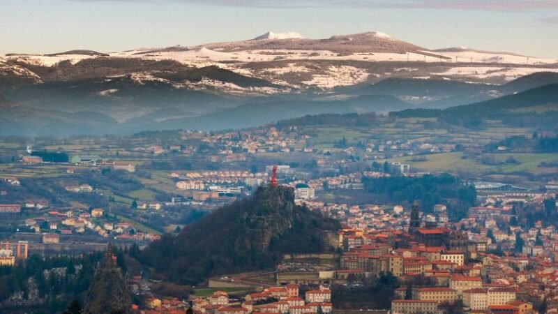 Vue panoramique d’une ville nichée dans une vallée, entourée de collines et de montagnes enneigées avec des monuments perchés sur des rochers.