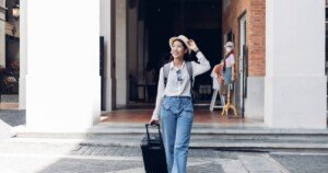 Voyageuse souriante tirant une valise dans une rue pavée, portant un chapeau et un sac à dos.