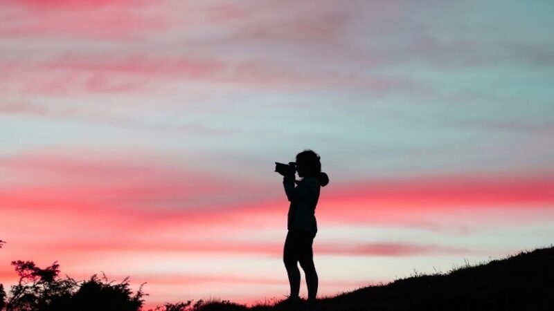 Silhouette d'une personne prenant une photo face à un ciel coloré rose et bleu au coucher du soleil.