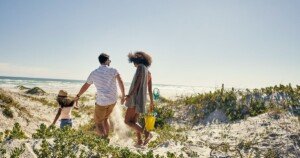 Famille marchant sur une plage de sable vers l’océan sous un ciel ensoleillé.