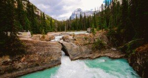 Vue spectaculaire d’une rivière turquoise traversant une formation rocheuse naturelle dans une forêt de sapins, avec des montagnes en arrière-plan.
