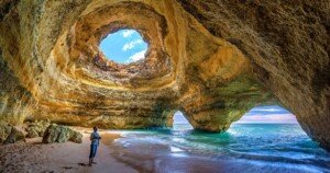 Touriste debout sur la plage intérieure de la grotte de Benagil, Algarve, Portugal, avec ses arches rocheuses et l’ouverture naturelle au plafond laissant passer la lumière.